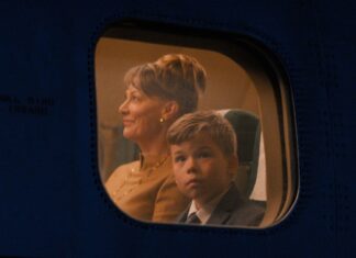 Woman with short hair and a young boy in a suit seated by an airplane window, illuminated by cabin light, looking ahead.