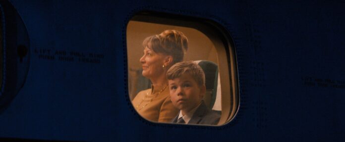 Woman with short hair and a young boy in a suit seated by an airplane window, illuminated by cabin light, looking ahead.