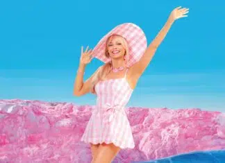 Woman in a pink gingham sundress and wide pink hat, arms raised, posing on a surreal pink rock landscape under a bright blue sky.