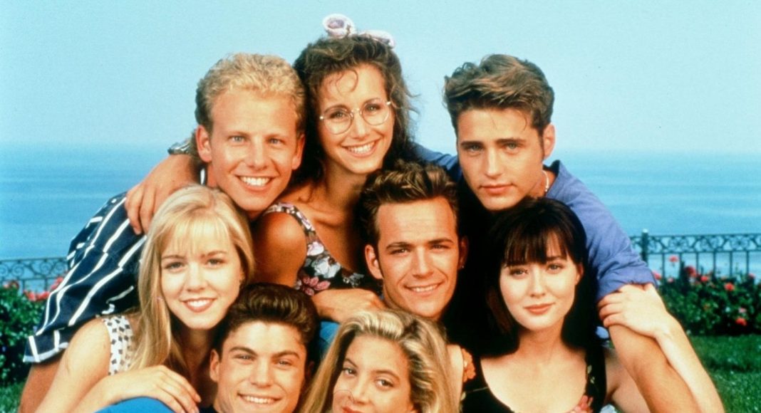 Group of eight young friends posing closely for a photo outdoors by the coast, all smiling under a bright blue sky.
