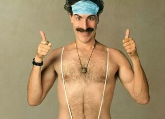 Sacha Baron Cohen não acha que vai voltar a viver o ‘Borat’ nos cinemas Smiling man with a blue surgical cap, shirtless, wearing suspenders and a necklace, showing thumbs up in a humorous medical-themed pose.