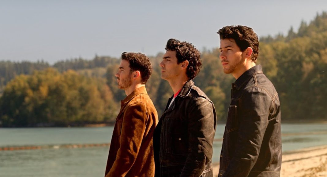 camp-rock-3-1280-x-720 Three men stand on a sandy lakeside, looking off into the distance, wearing casual jackets.