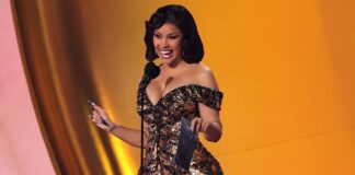 Woman in a sparkling off‑the‑shoulder gown smiles at a microphone on stage, holding a dark award plaque and a pen-like object.