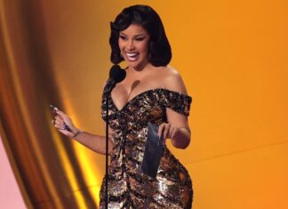 Woman in a sparkling off‑the‑shoulder gown smiles at a microphone on stage, holding a dark award plaque and a pen-like object.