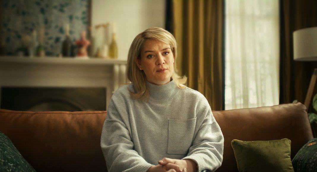 Blonde woman sits on a brown leather couch in a cozy living room, wearing a gray knit sweater and clasping her hands.