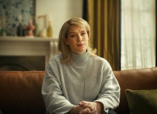 Crítica | ‘Casar com um Assassino?’ – Um dilema dilacerante marca novo TRUE CRIME da NETFLIX Blonde woman sits on a brown leather couch in a cozy living room, wearing a gray knit sweater and clasping her hands.