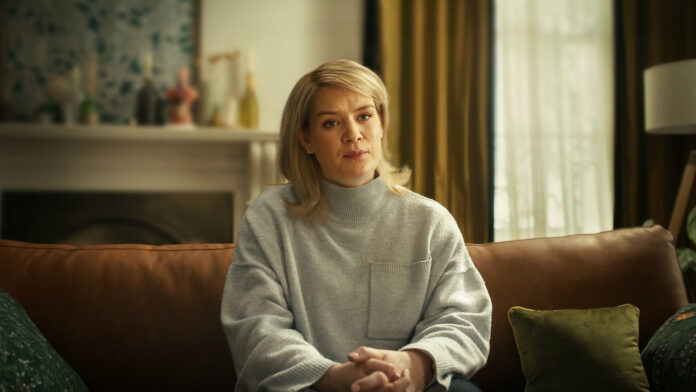 Blonde woman sits on a brown leather couch in a cozy living room, wearing a gray knit sweater and clasping her hands.
