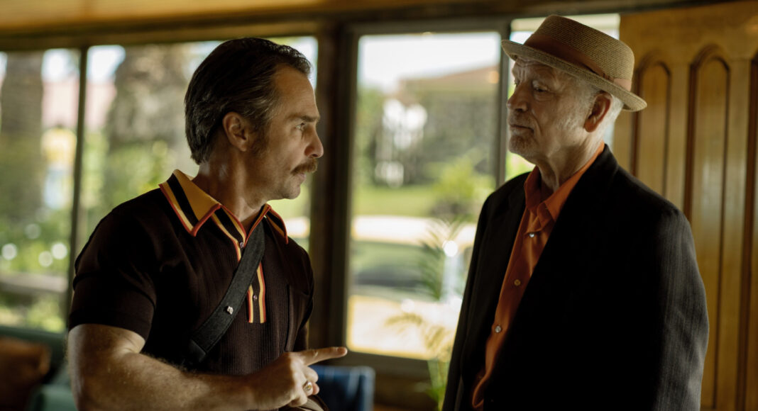 cavalo selvagem nove Two men converse indoors; the man on the left points as he speaks, wearing a dark polo with a crossbody strap.