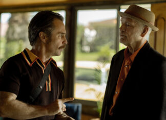 Two men converse indoors; the man on the left points as he speaks, wearing a dark polo with a crossbody strap.
