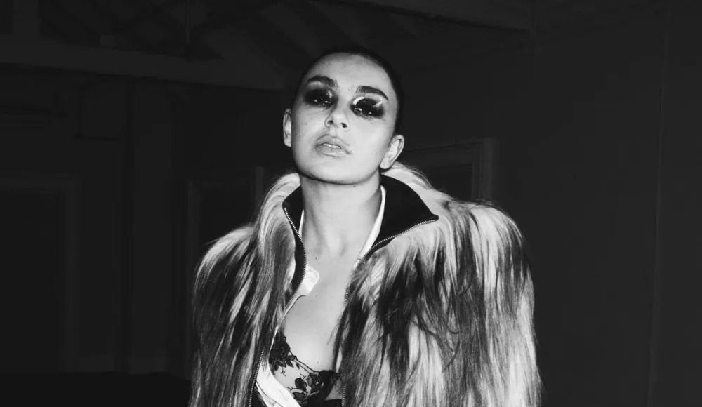 charli xcx Portrait of a woman wearing a long fur coat, with bold smoky makeup, looking slightly upward at the camera in a monochrome fashion shot.