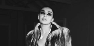 Portrait of a woman wearing a long fur coat, with bold smoky makeup, looking slightly upward at the camera in a monochrome fashion shot.