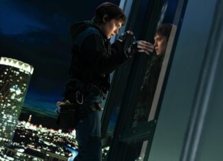 Climber in safety gear leaning against a glass wall at night, looking at their reflection with a lit city skyline below.