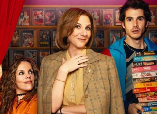 Promo still with three people; center woman in a beige plaid blazer smiles, flanked by a woman in orange and a man holding a tall pile of VHS tapes.