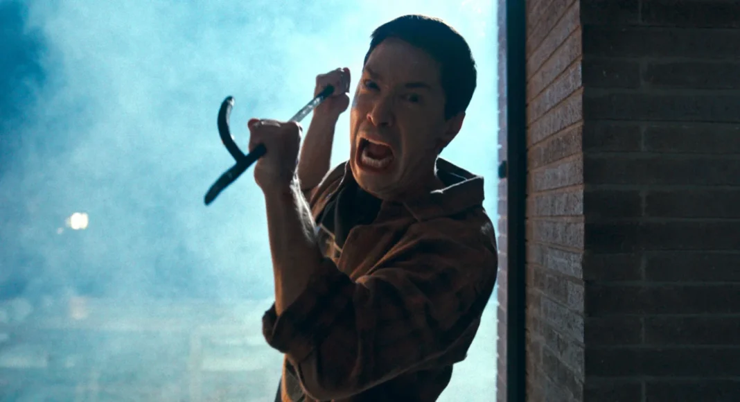 coyotes-JustinLong Man in a brown jacket screams while swinging a curved blade toward the camera in a smoky doorway with blue backlight.