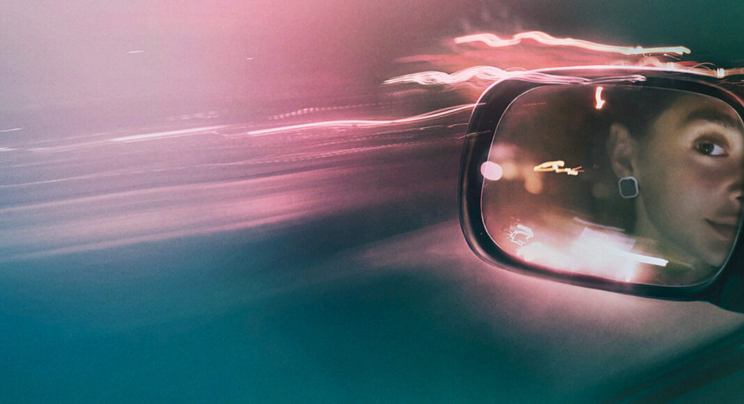 Side-view car mirror reflecting a person’s face at night with colorful light trails and motion blur in the background.
