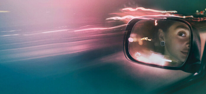 Side-view car mirror reflecting a person’s face at night with colorful light trails and motion blur in the background.