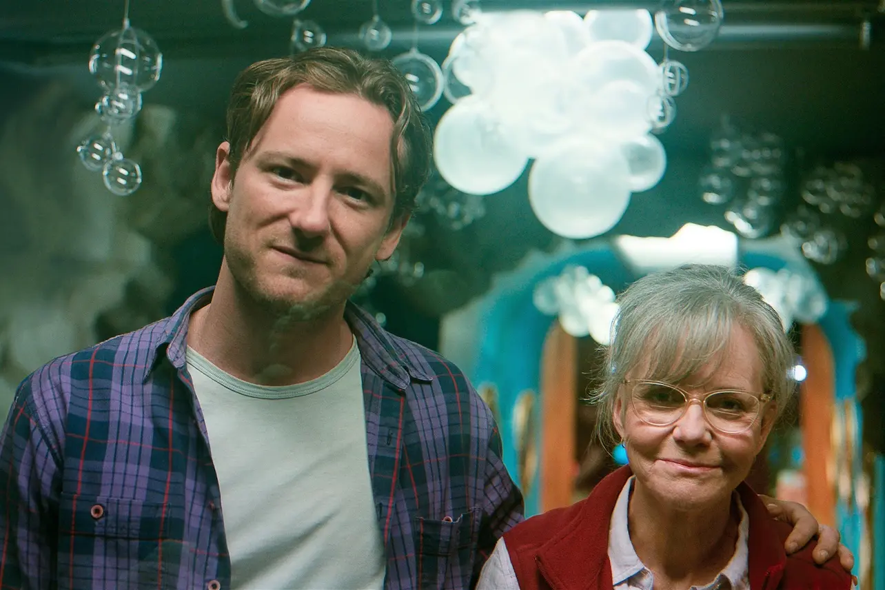Two people pose together, a man in a plaid shirt with arm around an older woman wearing glasses, smiling, with festive balloons in the background