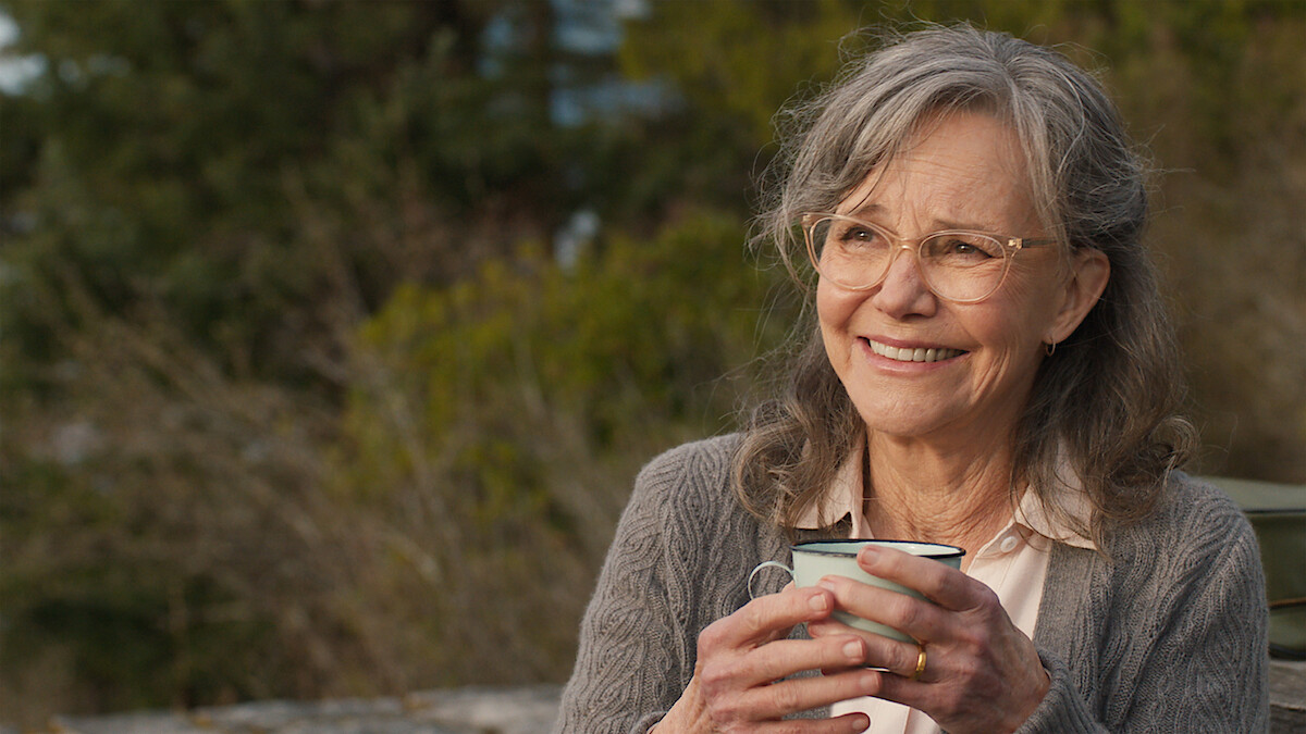 Older woman with gray hair and glasses smiling while holding a mug outdoors, wearing a gray cardigan.