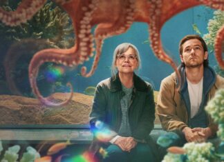 Two people sit and gaze up at a giant octopus above them in an aquarium exhibit, colorful coral around.