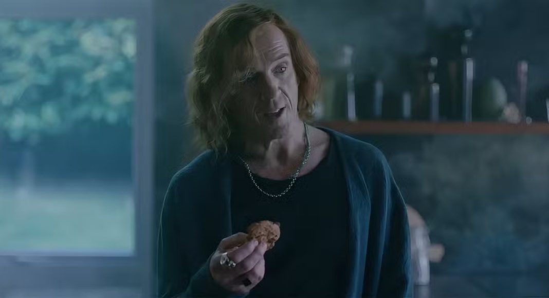 Man with shoulder-length hair in a dark blue cardigan holds a cookie while talking in a kitchen.