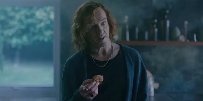 Man with shoulder-length hair in a dark blue cardigan holds a cookie while talking in a kitchen.