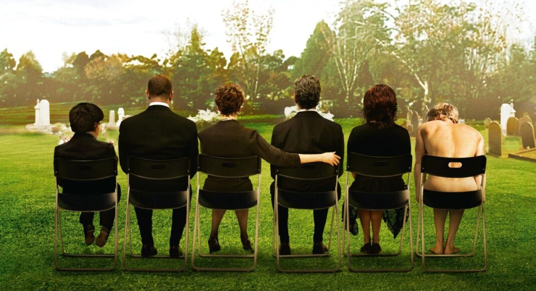 Six people sit in a row on folding chairs in a grassy cemetery, seen from behind, with tombstones in the background (somber gathering).