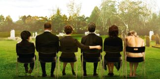 Six people sit in a row on folding chairs in a grassy cemetery, seen from behind, with tombstones in the background (somber gathering).