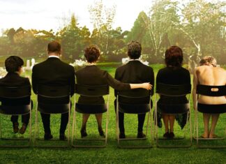 Tá sem dinheiro pra balada? 10 filmes que vão te fazer amar ficar em casa Six people sit in a row on folding chairs in a grassy cemetery, seen from behind, with tombstones in the background (somber gathering).