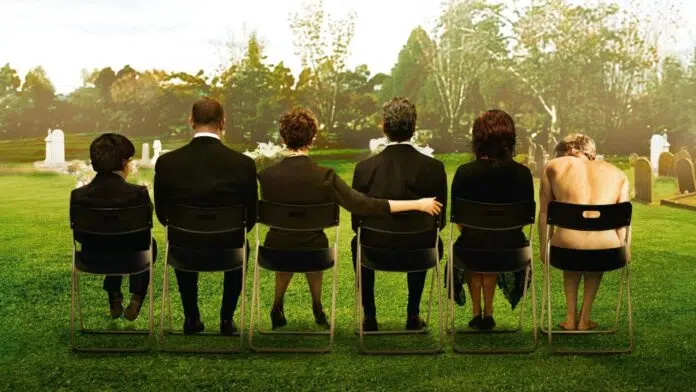 death Six people sit in a row on folding chairs in a grassy cemetery, seen from behind, with tombstones in the background (somber gathering).