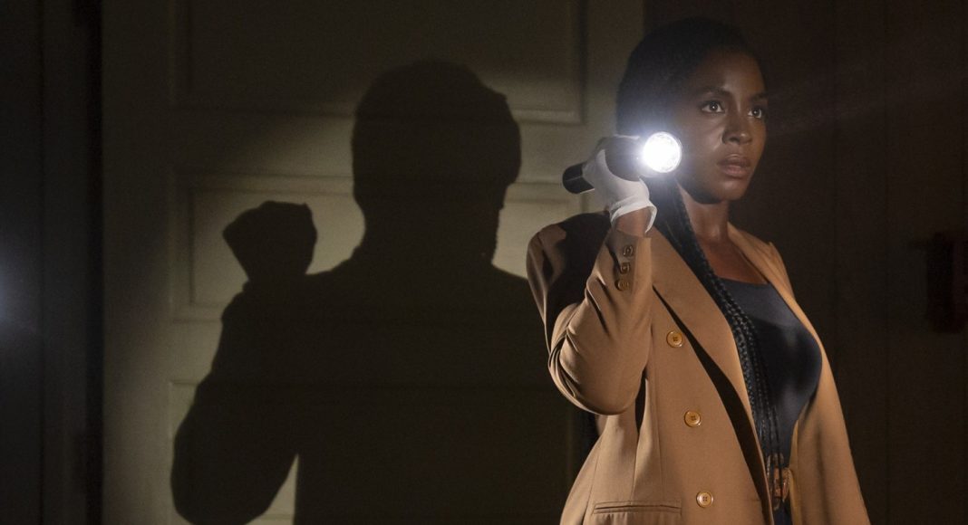 Woman in a tan blazer uses a flashlight in a dark hallway, creating a large shadow on the wall behind her.