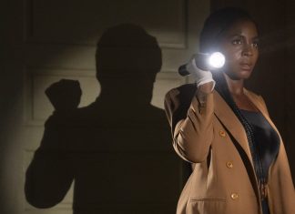 Woman in a tan blazer uses a flashlight in a dark hallway, creating a large shadow on the wall behind her.