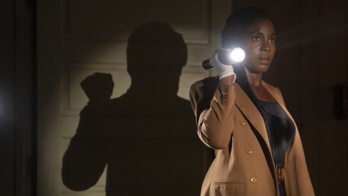 Woman in a tan blazer uses a flashlight in a dark hallway, creating a large shadow on the wall behind her.