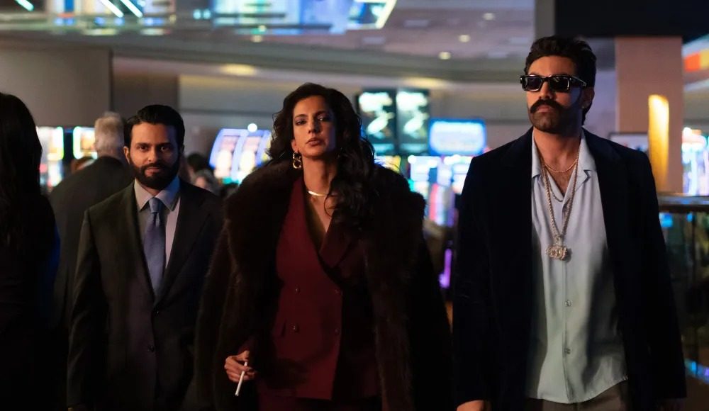 Three stylish adults walk through a bright casino lobby; a woman in a burgundy suit and fur coat leads, flanked by two men in suits.