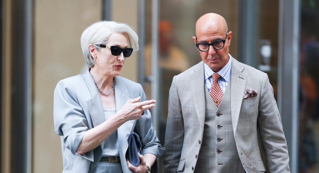 Two stylish older adults in light gray suits walk and chat on a city sidewalk while wearing sunglasses.