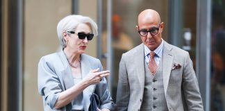 Two stylish older adults in light gray suits walk and chat on a city sidewalk while wearing sunglasses.