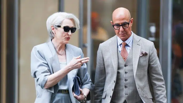 Two stylish older adults in light gray suits walk and chat on a city sidewalk while wearing sunglasses.