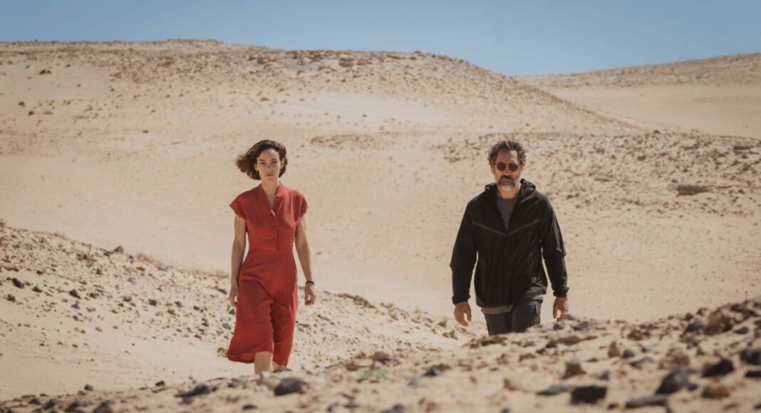 Two adults walk through a barren desert with rolling sand dunes and a clear blue sky behind them, one in red and the other in dark clothing on a sunny day.