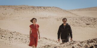 Two adults walk through a barren desert with rolling sand dunes and a clear blue sky behind them, one in red and the other in dark clothing on a sunny day.