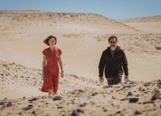 Two adults walk through a barren desert with rolling sand dunes and a clear blue sky behind them, one in red and the other in dark clothing on a sunny day.