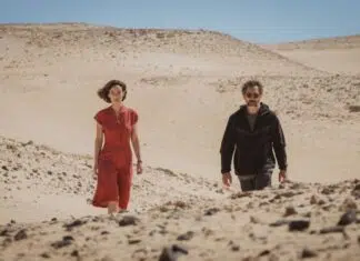 Two adults walk through a barren desert with rolling sand dunes and a clear blue sky behind them, one in red and the other in dark clothing on a sunny day.