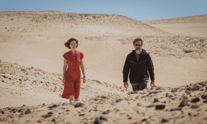 Two adults walk through a barren desert with rolling sand dunes and a clear blue sky behind them, one in red and the other in dark clothing on a sunny day.