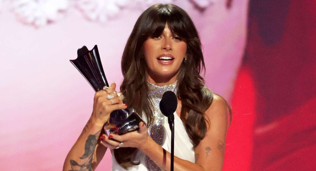 ella langley Woman on stage holding a shiny trophy and speaking into a microphone, with a pink-red backdrop behind her.