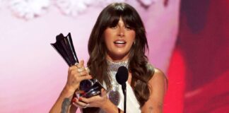 Woman on stage holding a shiny trophy and speaking into a microphone, with a pink-red backdrop behind her.