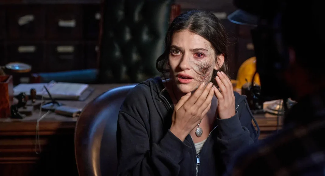 ellarubin-untildawn Woman with facial prosthetic scar touching her cheek in a dim, cluttered office.