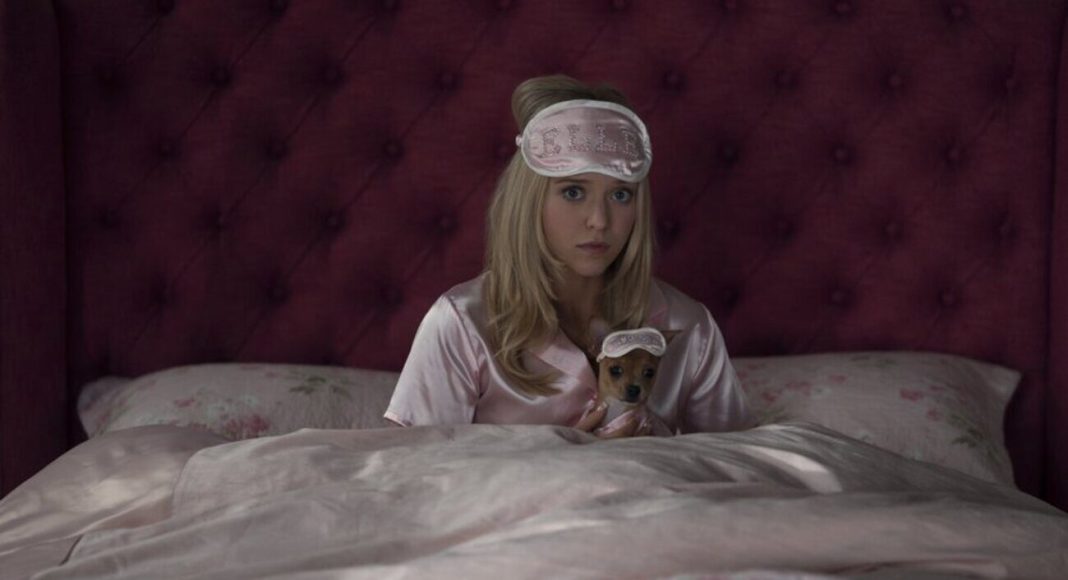 Woman in pink satin pajamas sits up in bed, wearing a blush sleep mask, holding a tiny dog in a matching sleep mask.