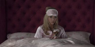 Woman in pink satin pajamas sits up in bed, wearing a blush sleep mask, holding a tiny dog in a matching sleep mask.