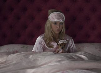 Woman in pink satin pajamas sits up in bed, wearing a blush sleep mask, holding a tiny dog in a matching sleep mask.