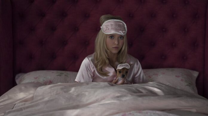 Woman in pink satin pajamas sits up in bed, wearing a blush sleep mask, holding a tiny dog in a matching sleep mask.