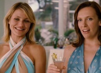 Two women in halter-style dresses smile at the camera, each holding a cocktail at a bright indoor party set near a terrace.