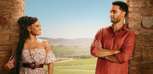 Woman in a floral off-shoulder dress with a belt smiles at a man in a red shirt as they stand between stone pillars with a rural landscape behind them.